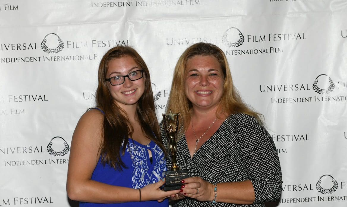 Two women holding a trophy in front of a white wall.