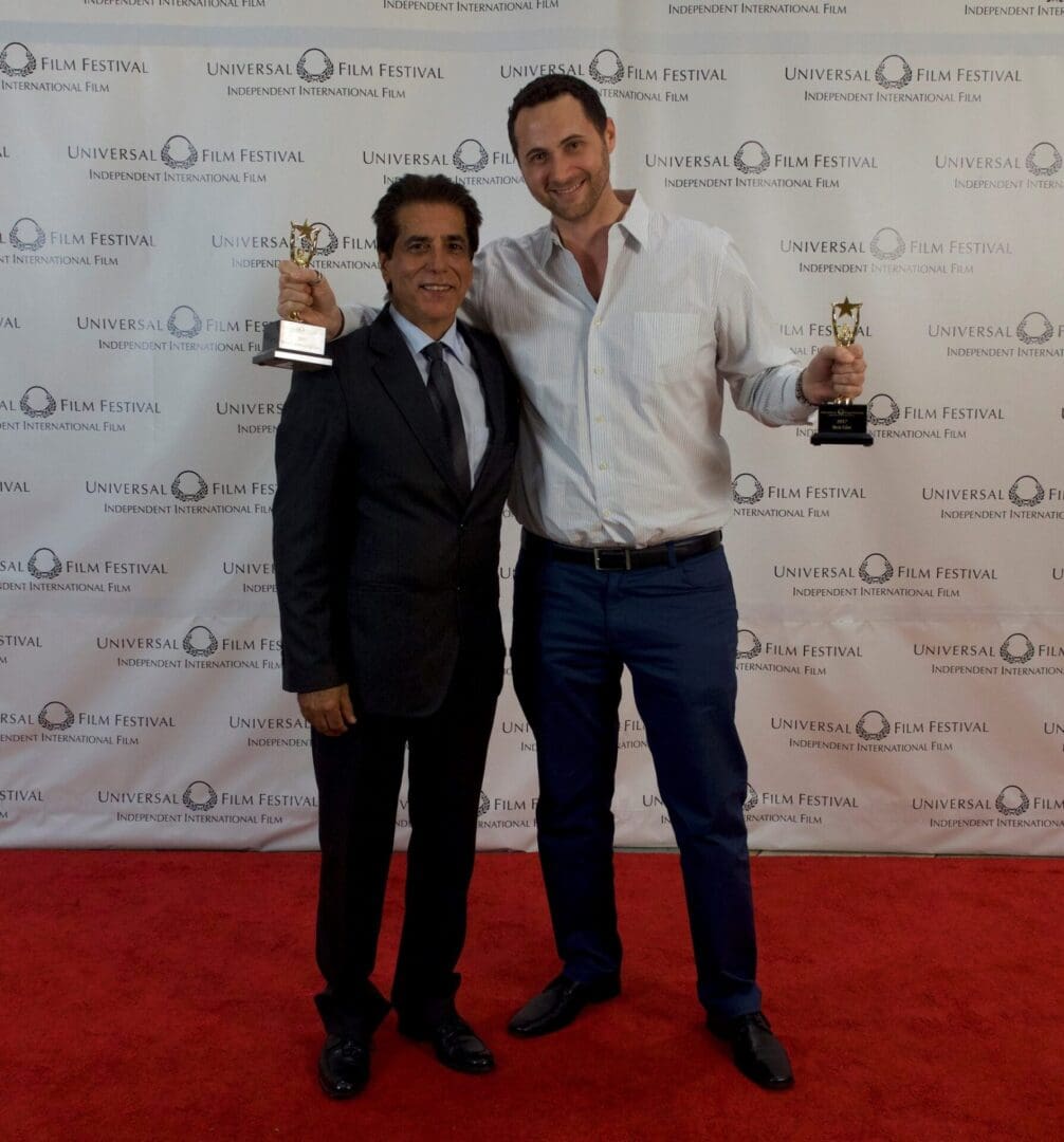 Two men holding up their awards on a red carpet.